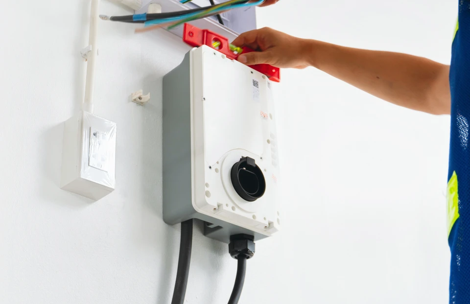 Photographie réaliste d'un électricien professionnel effectuant l'installation d'une borne de recharge pour véhicule électrique sur la façade extérieure d'une maison résidentielle ou d'un immeuble tertiaire. L'électricien porte des équipements de sécurité (gilet, outils professionnels), travaille méthodiquement sur le câblage et le branchement de la borne. Le contexte montre clairement une installation complète et soignée, représentative de services de mobilité électrique professionnels et certifiés IRVE.